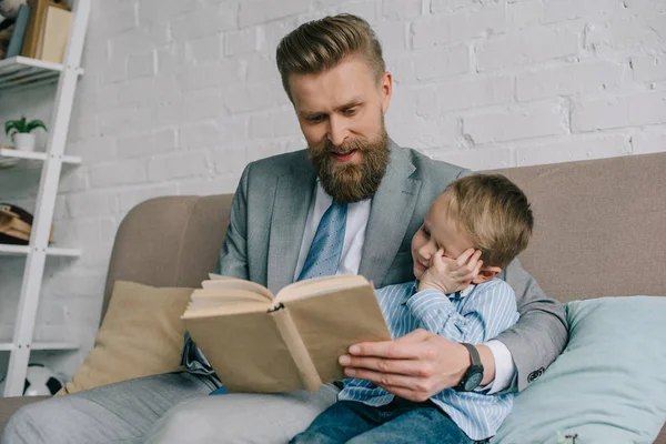 Businessman and little son reading book together an sofa at home, work and life balance — Stock Photo