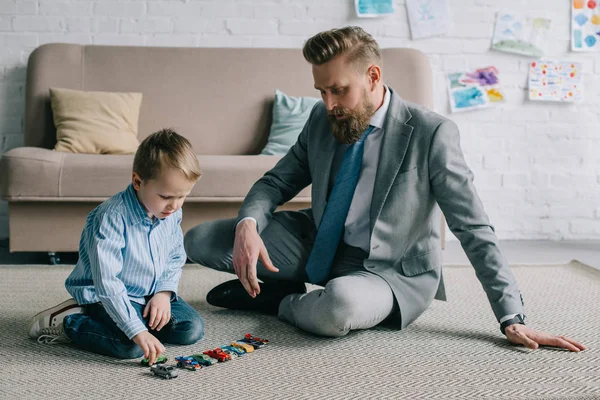 Homem de negócios em terno e pequeno filho brincando com carros de brinquedo no chão em casa, trabalho e conceito de equilíbrio de vida — Fotografia de Stock