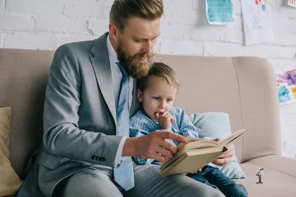 Businessman and little son reading book together an sofa at home, work and life balance — Stock Photo