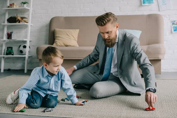 Homem de negócios em terno e pequeno filho brincando com carros de brinquedo no chão em casa, trabalho e conceito de equilíbrio de vida — Fotografia de Stock