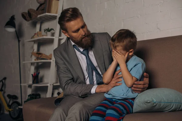 Homem em terno de negócios animando filho chateado no sofá em casa, trabalho e conceito de equilíbrio de vida — Fotografia de Stock
