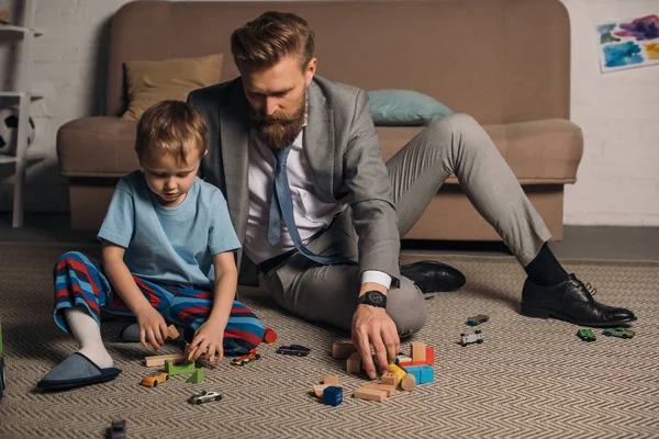 Hombre de negocios en traje y pequeño hijo jugando con bloques de madera en el suelo en el hogar, el trabajo y el concepto de equilibrio de la vida - foto de stock