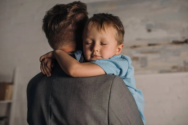 Niño pequeño con los ojos cerrados abrazando padre en casa - foto de stock
