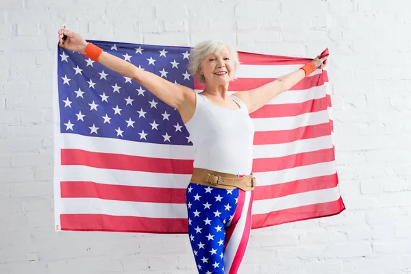 Heureuse femme âgée en vêtements de sport tenant drapeau américain et souriant à la caméra — Photo de stock
