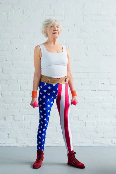 Полный вид пожилой женщины в патриотической спортивной одежде, держащей гантели и смотрящей в камеру — стоковое фото