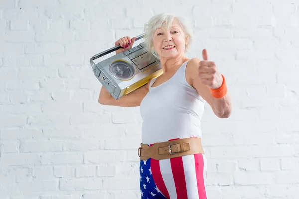 Happy senior woman in sportswear holding tape recorder and showing thumb up — Stock Photo