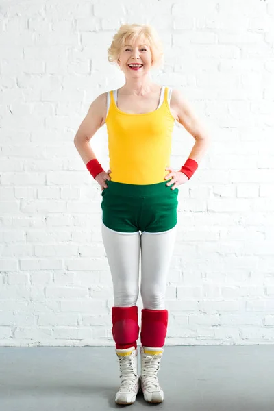 Feliz mujer mayor en ropa deportiva de pie con las manos en la cintura y sonriendo a la cámara — Stock Photo