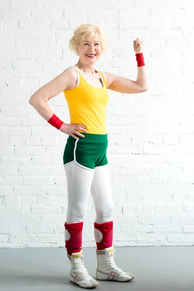 Happy senior woman in sportswear showing biceps and smiling at camera — Stock Photo