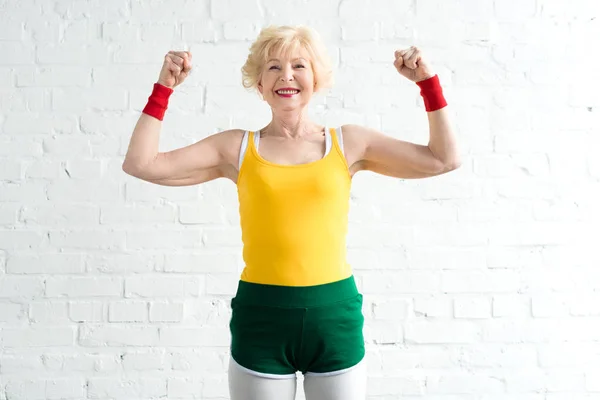 Feliz mujer mayor en ropa deportiva mostrando los músculos y sonriendo a la cámara - foto de stock