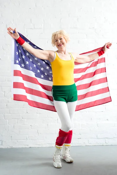 Full length view of happy senior sportswoman holding us flag and smiling at camera — Stock Photo