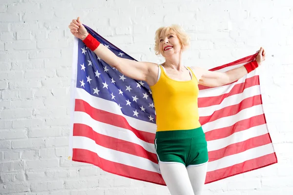 Alegre deportivo senior mujer en ropa deportiva sosteniendo nosotros bandera y mirando hacia otro lado - foto de stock