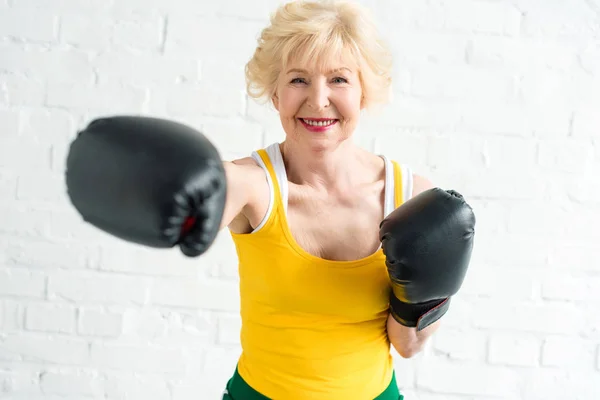 Feliz mujer mayor en el entrenamiento de guantes de boxeo y sonriendo a la cámara - foto de stock