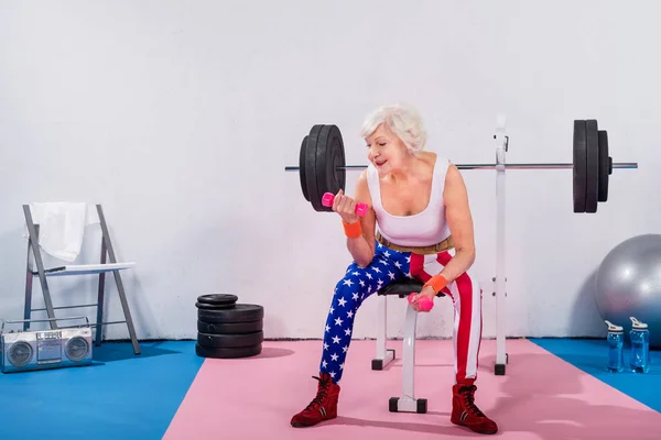 Smiling senior woman in sportswear sitting and training with dumbbells in gym — Stock Photo