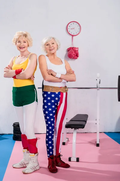 Donne anziane sportive in piedi con le braccia incrociate e sorridenti alla fotocamera in palestra — Foto stock