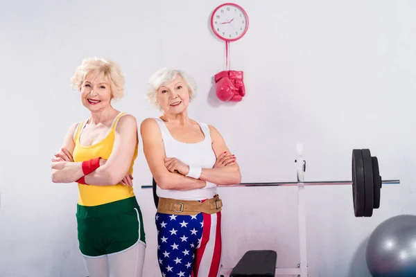 Sportifs seniors debout avec les bras croisés et souriant à la caméra dans la salle de gym — Photo de stock
