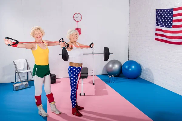 Happy senior sportswomen training with resistance bands and smiling at camera in gym — Stock Photo