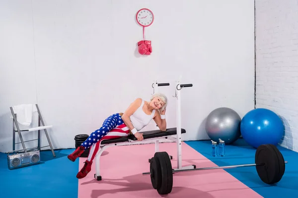 Felice sportiva anziana sdraiata sulla panchina e sorridente alla macchina fotografica in palestra — Foto stock