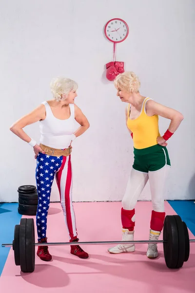 Старшие спортсменки, стоящие с руками на талии и улыбающиеся друг другу в спортзале — стоковое фото