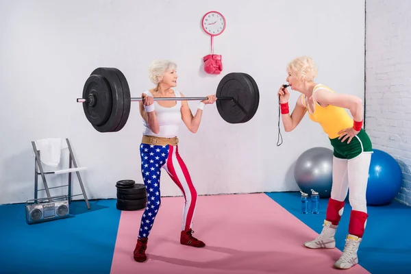 Senior female coach with whistle looking at senior sportswoman lifting barbell in gym — Stock Photo