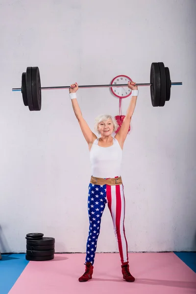 Sportive senior woman in patriotic sportswear holding barbell and smiling at camera — Stock Photo