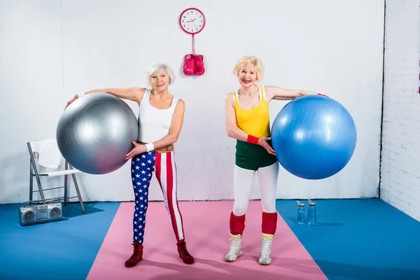 Deportivo señoras mayores sosteniendo bolas de fitness y sonriendo a la cámara en el gimnasio - foto de stock