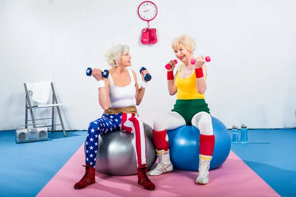 Mulheres idosas em treinamento sportswear com halteres enquanto sentado em bolas de fitness e sorrindo uns aos outros — Fotografia de Stock