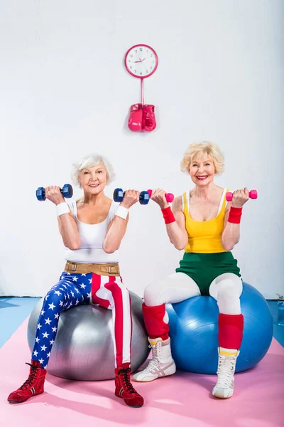 Mujeres mayores en entrenamiento de ropa deportiva con pesas mientras están sentadas en bolas de fitness y sonriendo a la cámara - foto de stock