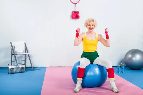 Femme âgée sportive souriante assise sur un ballon de fitness et faisant de l'exercice avec des haltères — Photo de stock