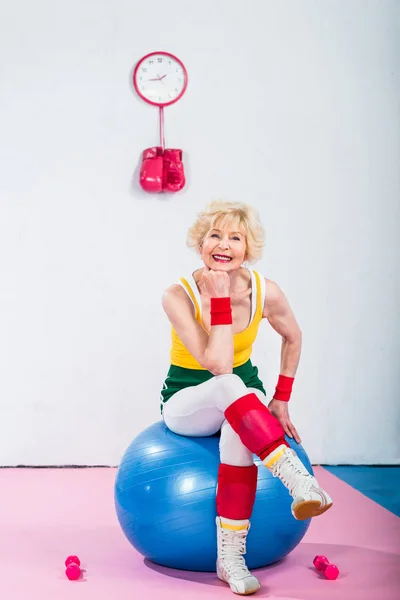 Feliz deportivo senior mujer sentado en fitness pelota y sonriendo a la cámara - foto de stock