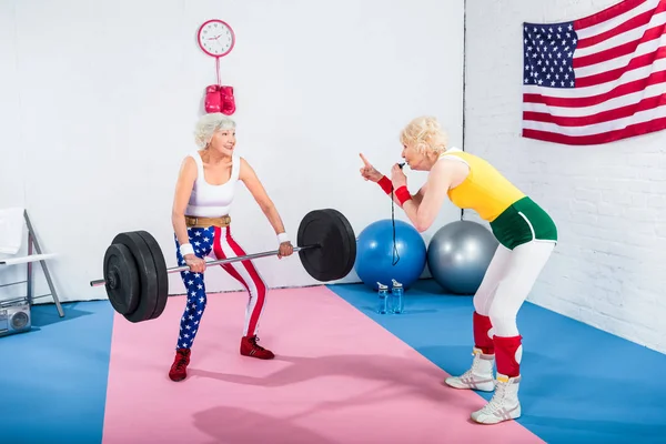 Desportivo sênior mulher soprando assobio e olhando para idosos desportista levantar barbell no ginásio — Fotografia de Stock