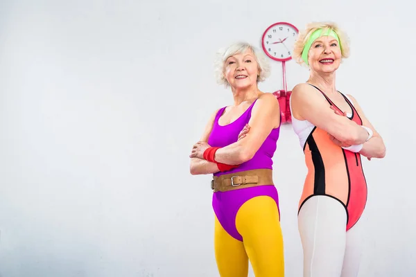 Low angle view of happy senior sportswomen standing with crossed arms and smiling at camera — Stock Photo