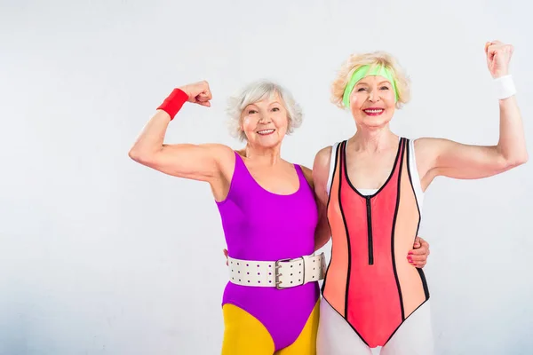 Happy senior sportswomen showing muscles and smiling at camera isolated on grey — Stock Photo