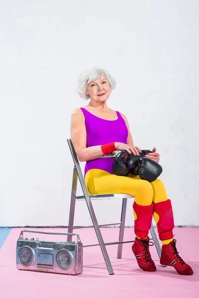 Schöne sportliche Seniorin mit Boxhandschuhen sitzend und lächelnd in die Kamera — Stock Photo