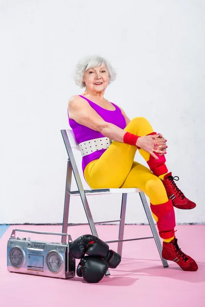 Happy senior sportswoman sitting near tape recorder with boxing gloves and smiling at camera — Stock Photo