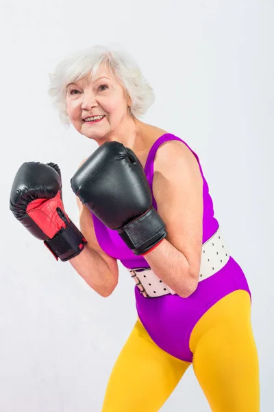 Ritratto di bella signora anziana sportiva in guanti da boxe sorridente alla fotocamera isolata sul grigio — Foto stock