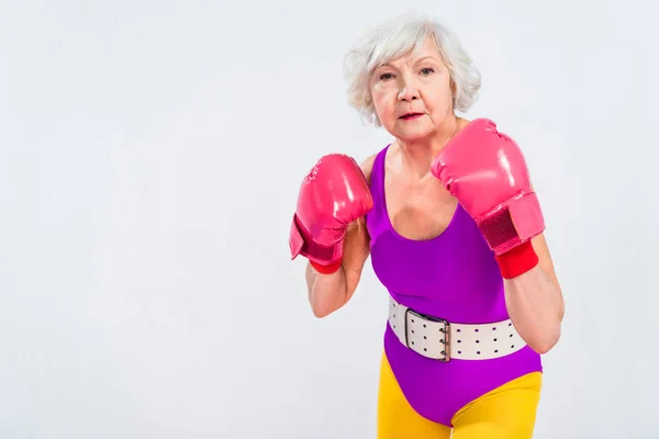 Mujer mayor en ropa deportiva y guantes de boxeo mirando a la cámara aislada en gris - foto de stock