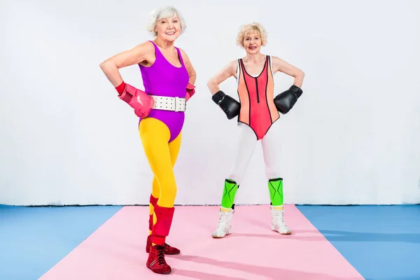 Vista completa de las señoras mayores deportivas en guantes de boxeo de pie con las manos en la cintura y sonriendo a la cámara - foto de stock