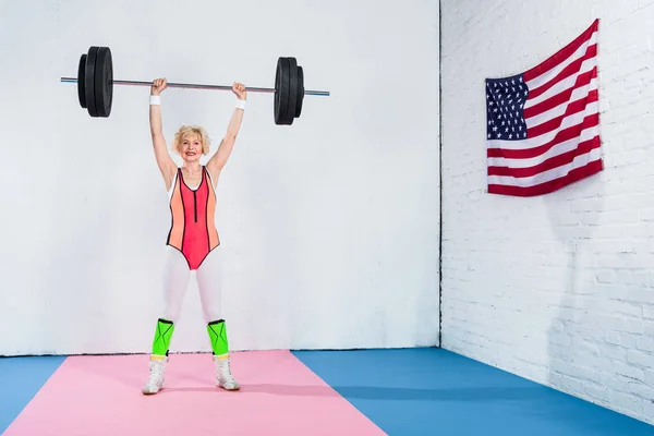 Pleine longueur vue de sportif senior femme levant haltère et souriant à la caméra — Photo de stock