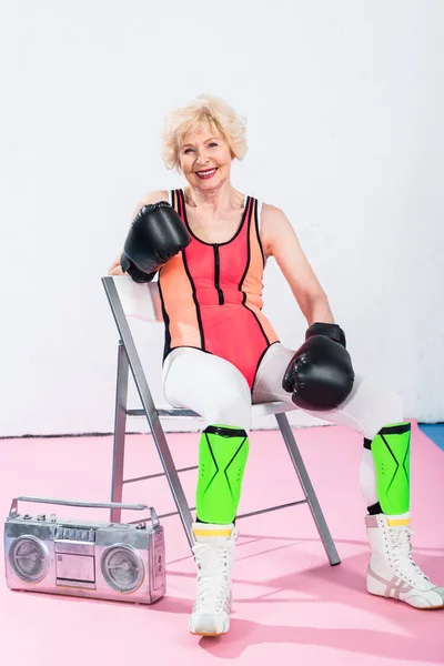 Sportliche Seniorin in Boxhandschuhen sitzt mit Tonbandgerät und lächelt in die Kamera — Stockfoto