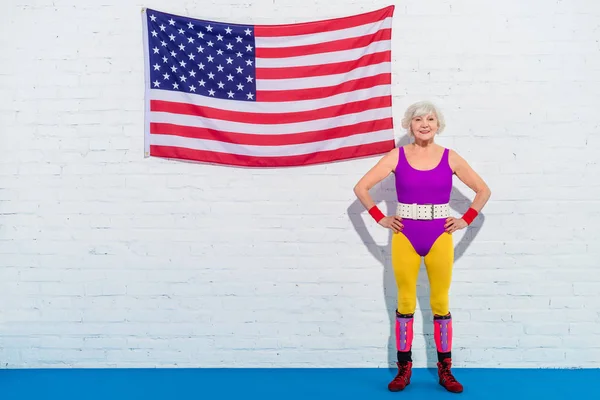 Happy senior woman standing with hands on waist near us flag and smiling at camera — Stock Photo