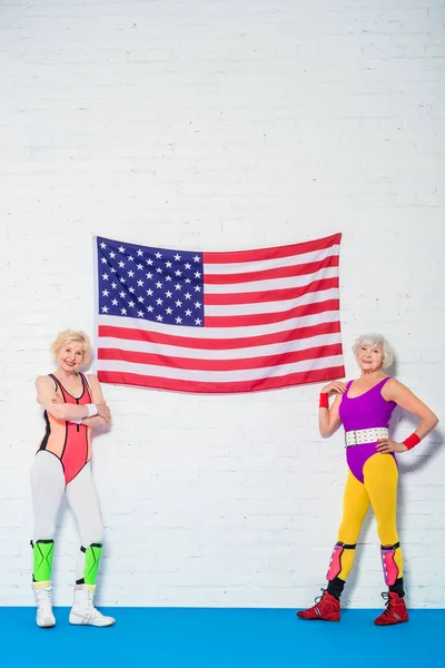 Ganzkörperansicht schöner älterer Sportlerinnen, die neben der amerikanischen Flagge stehen und in die Kamera lächeln — Stockfoto