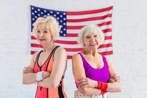 Belles femmes âgées sportives debout avec les bras croisés et souriant à la caméra contre le drapeau américain — Photo de stock