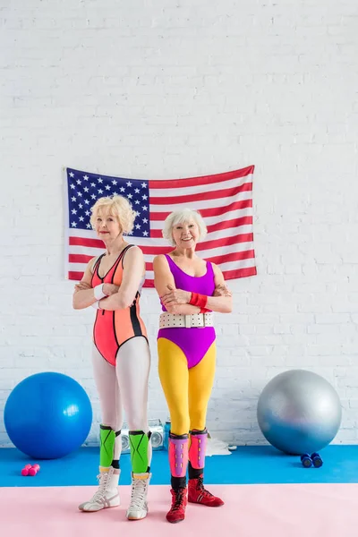 Впевнені старші спортсмени стоять з схрещеними руками і посміхаються на камеру в спортзалі з американським прапором — стокове фото