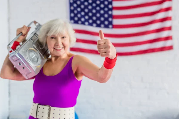 Happy senior woman in sportswear holding tape recorder and showing thumb up — Stock Photo