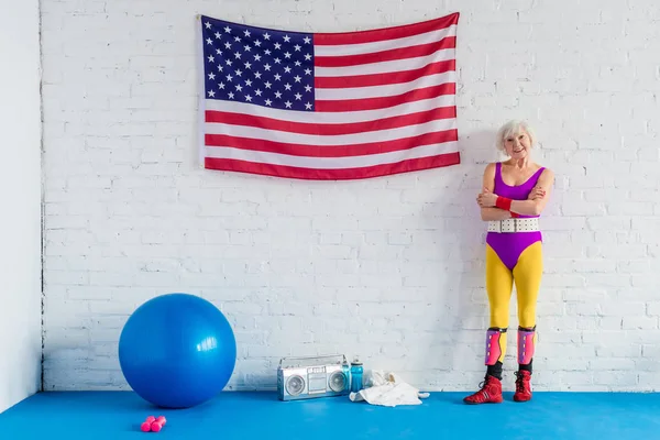 Красивая старшая спортсменка, стоящая со скрещенными руками возле американского флага и улыбающаяся в камеру — стоковое фото