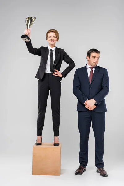 Femme d'affaires heureuse avec coupe trophée debout sur le podium, collègue bouleversé debout près, isolé sur gris — Photo de stock