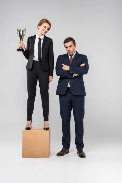 Felice donna d'affari con coppa trofeo in piedi sul podio, uomo d'affari sconvolto in piedi vicino, isolato su grigio — Foto stock