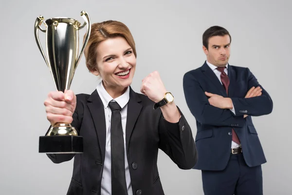 Excited businesswoman with trophy cup and upset businessman behind, isolated on grey — Stock Photo