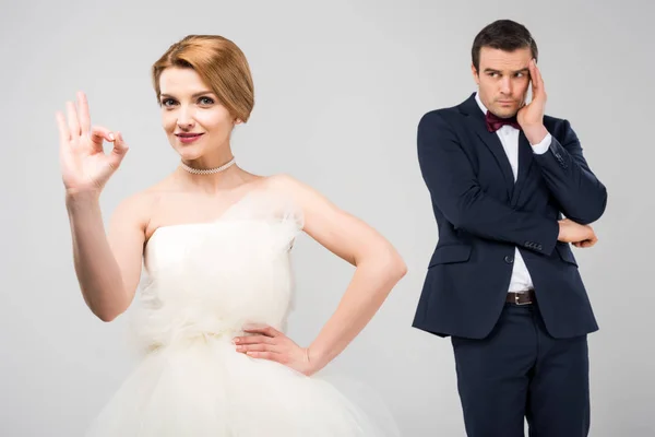 Braut im Brautkleid zeigt Ok-Zeichen, während besorgter Bräutigam dahintersteht, isoliert auf grau, Feminismus-Konzept — Stockfoto