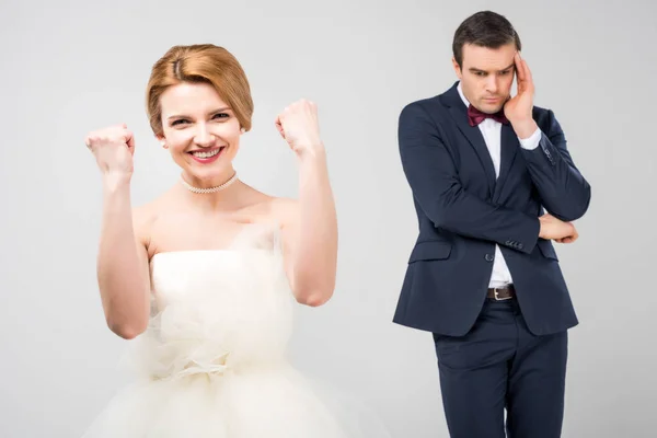 Excited bride in wedding dress and worried groom behind, isolated on grey, feminism concept — Stock Photo